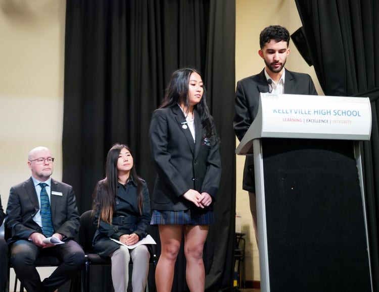 Two students standing at lectern addressign an audience. 2 teachers seated behind them.